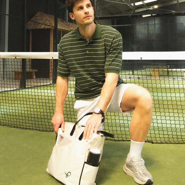 A man in a green and white striped polo shirt, white shorts, and athletic shoes kneels on a padel court holding a white padel bag with black accents.